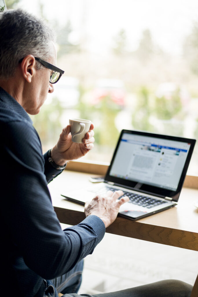 Elderly businessmandrinking coffee while responding to comments on a laptop.